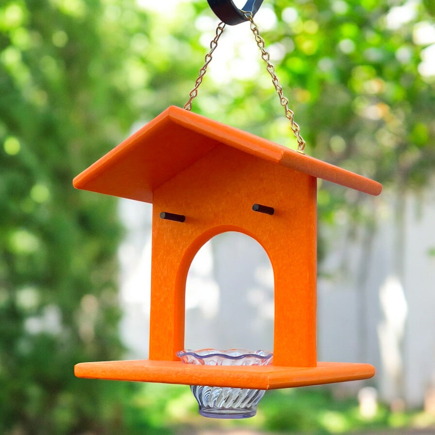 An orange Oriole bird feeder with a plastic jelly cup hangs from a metal chain, with trees in the background.